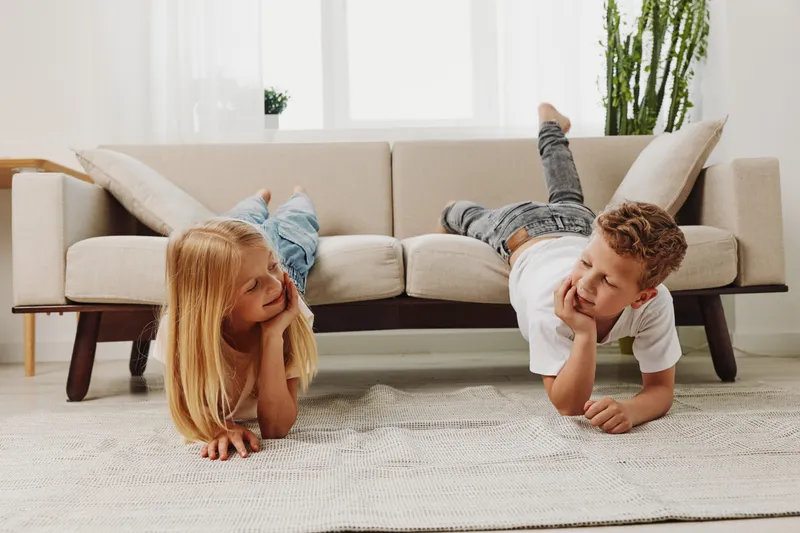 Two playful children lying on the floor in front of a beige sofa in a bright living room. The boy and girl smile while relaxing, creating a warm and friendly family atmosphere.