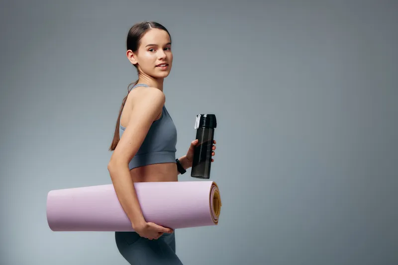Active woman in athletic wear holding a yoga mat and water bottle, ready for a workout in a clean studio backdrop, showcasing fitness and readiness.