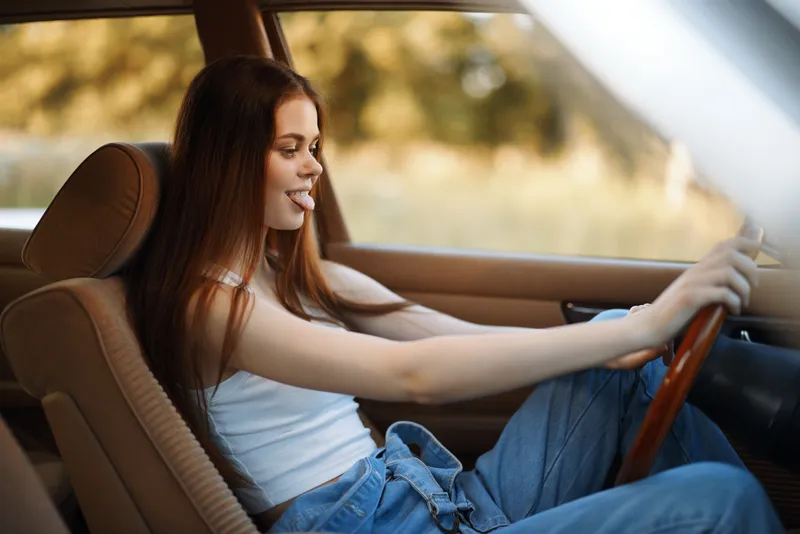 Young woman smiling and enjoying a road trip in a vintage car, wearing casual denim attire and posing playfully with a joyful expression.
