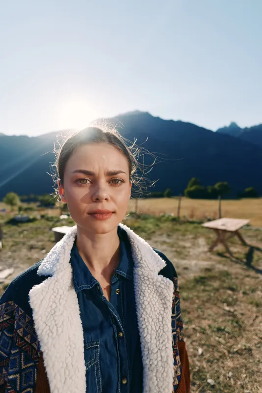 Woman portrait outdoors in mountains with sunlight behind her, young person wearing denim shirt and shearling jacket in field, natural expression, travel lifestyle and seasonal fashion.