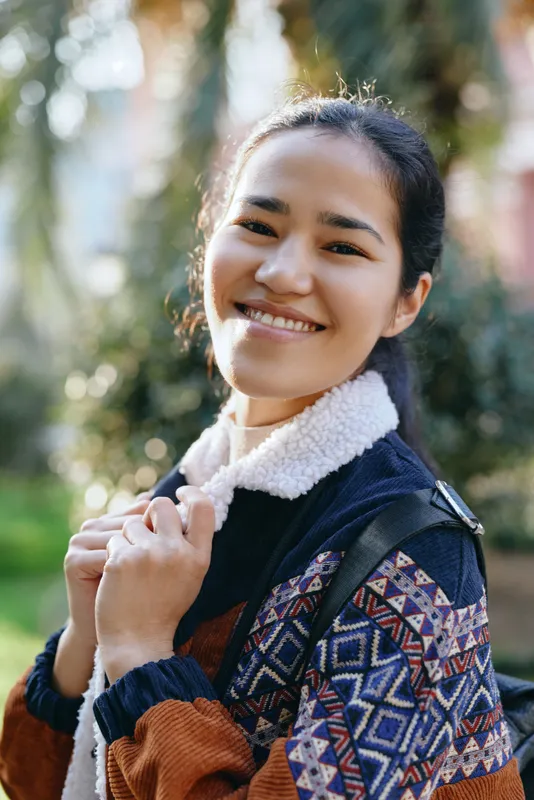 Smiling woman outdoors in a patterned jacket with a warm fleece collar, autumn sunlight streaming through trees, casual fashion and cheerful lifestyle moment