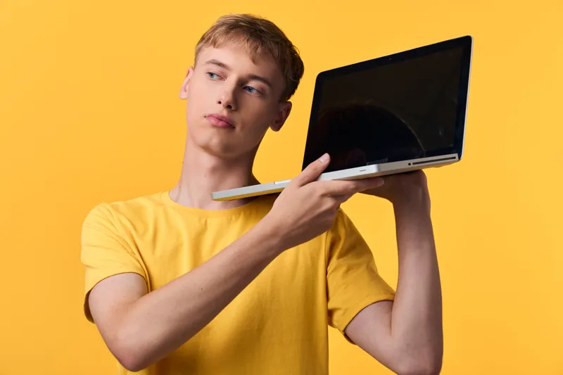 Man holding a laptop at shoulder against a bright yellow background, showcasing casual tech style, portable device, modern screen, and flexible workstation concept for lifestyle innovation and