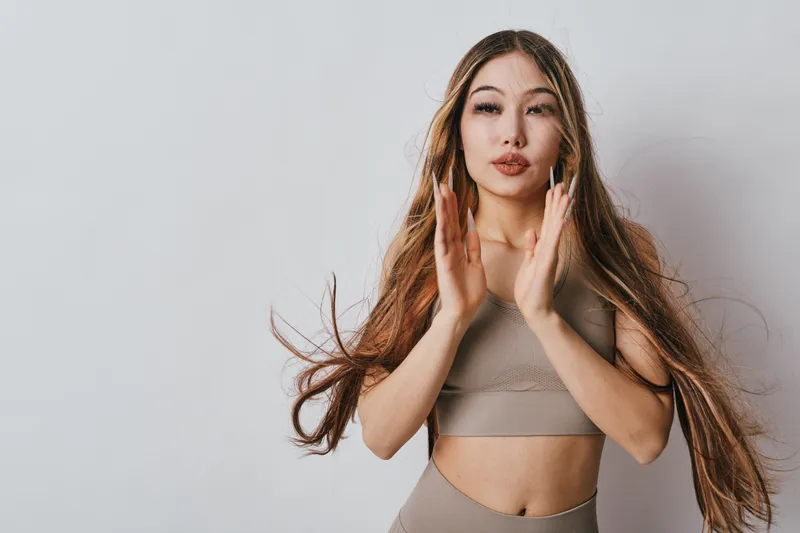 Young woman with long hair in sportswear posing with hands raised during fitness workout indoor. Female athlete, training, health and active lifestyle concept in studio.