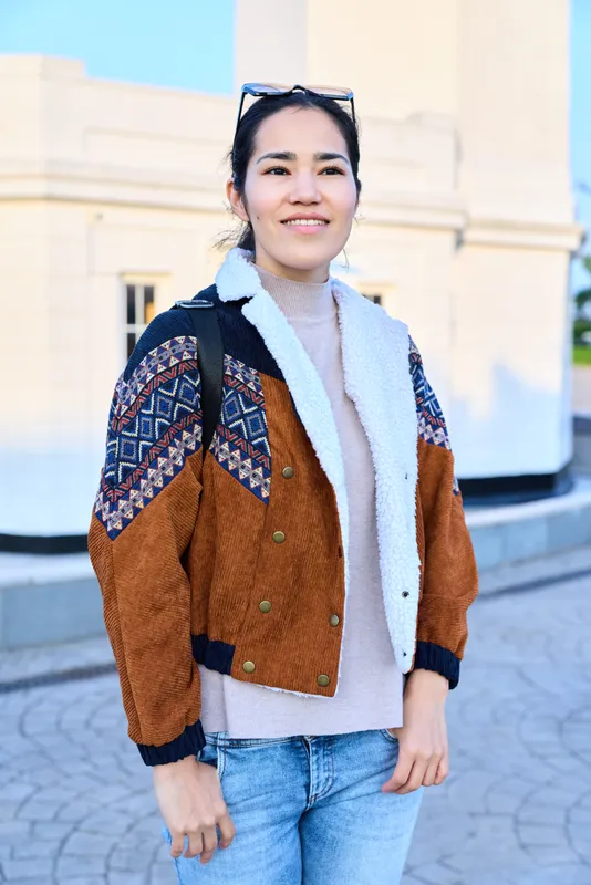 Woman wearing a brown and blue patterned jacket with shearling collar, light knit sweater and jeans, sunglasses on head, standing in a sunny urban plaza, showcasing casual street fashion and a