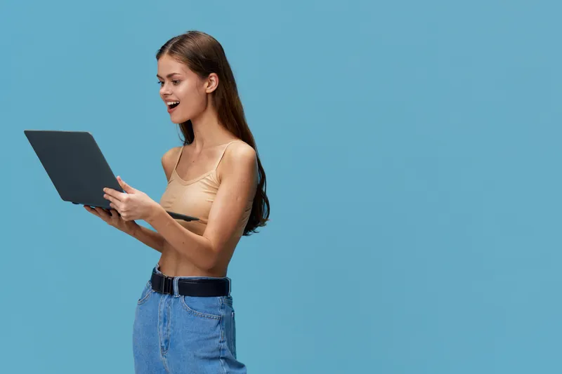 young woman with laptop smiling and standing against blue background wearing casual beige top and jeans, expressing excitement and focus in natural light