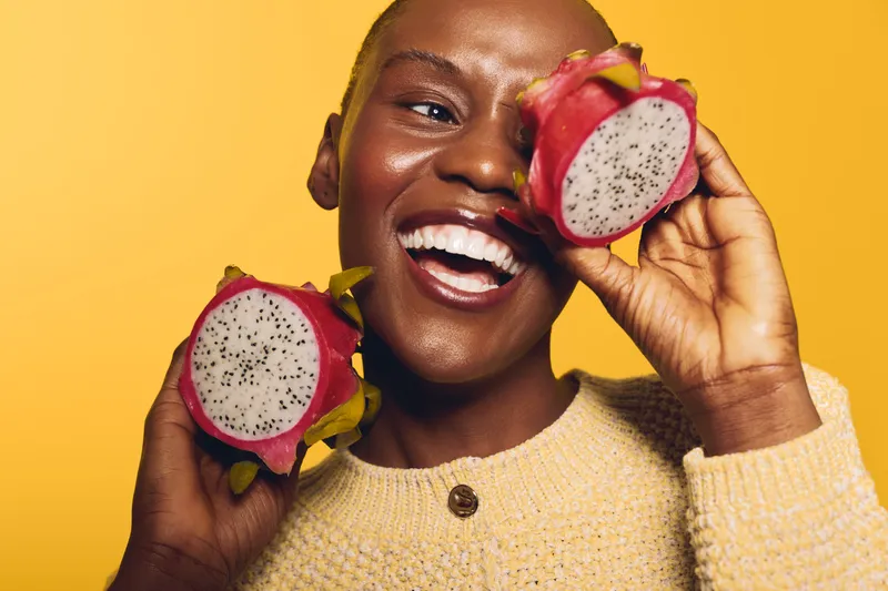 Happy African woman with dark skin holds two halves of dragon fruit near face, smiling brightly on yellow background. Concept of healthy lifestyle, tropical fruit, and joyful mood.