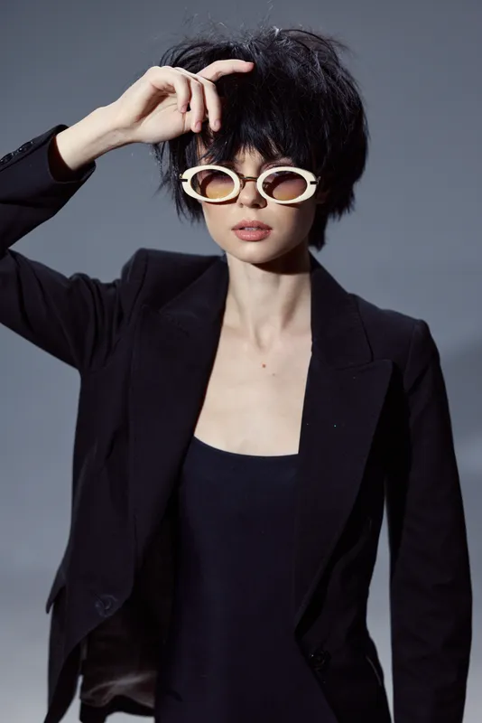 Stylish woman with short black hair wearing trendy sunglasses and a black blazer posing confidently in studio light.