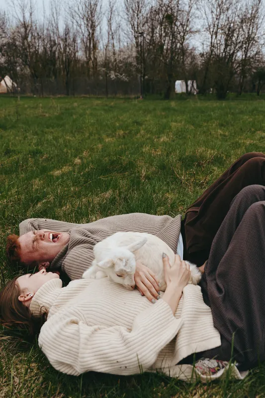 A young couple lies on the grass in casual sweaters holding a white baby goat, enjoying a relaxed lifestyle moment outdoors in early spring.