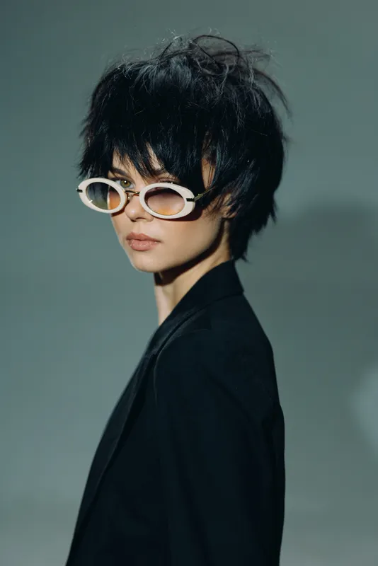 Stylish woman wearing unique sunglasses with short dark hair and a black outfit, posing confidently against a muted background.