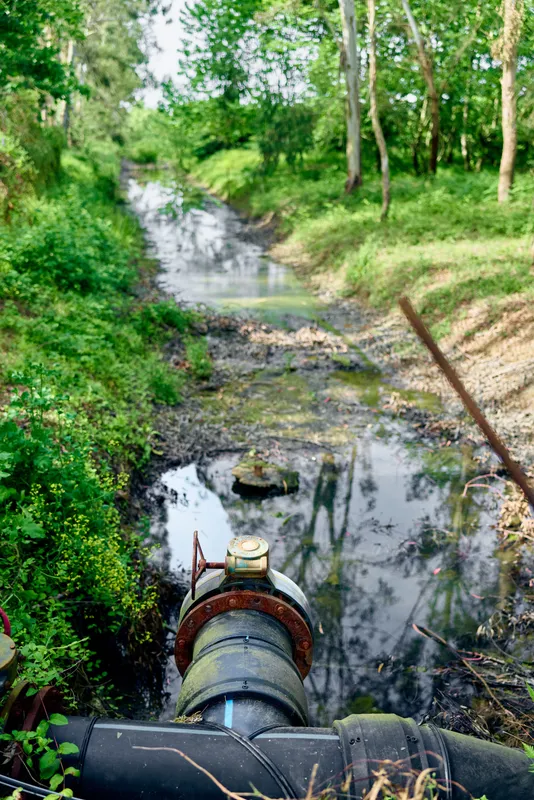 Pipe leaking water into a shallow stream in a forest, rusty valve and visible rust stains, signs of pollution and erosion along the creek and small riverbank in rural landscape.