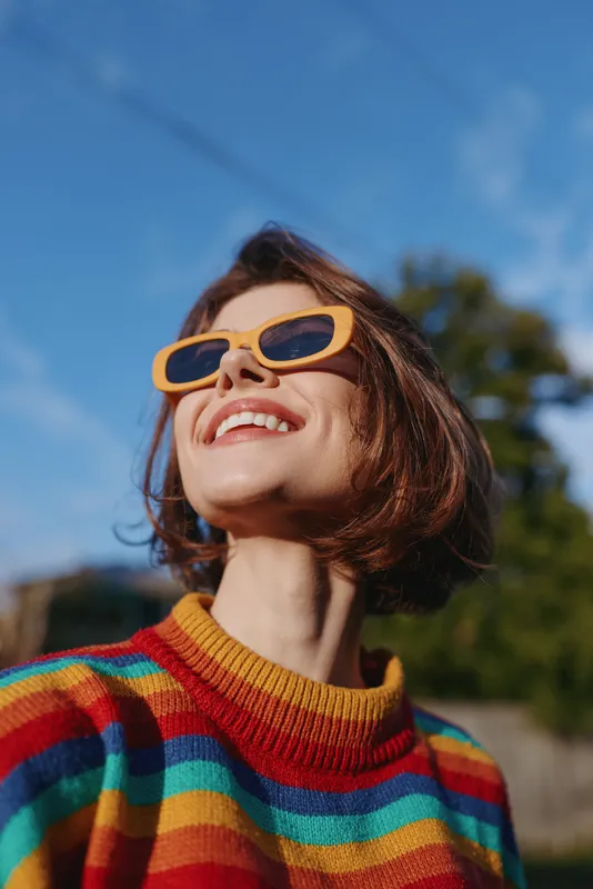 Woman short hair smile sunglasses rainbow sweater portrait outdoors happy natural light, colorful fashion closeup in sunny blue sky, joyful relaxed lifestyle and candid expression.