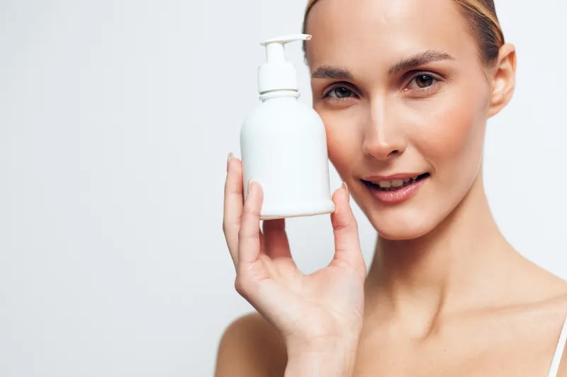 A young woman with clear skin holds a white cosmetic bottle near her face, smiling softly against a plain white background. Concept for skincare, beauty, and natural care.