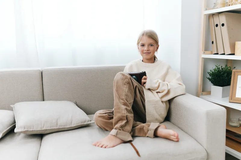 Young girl sits on a light gray sofa, holding a tablet, relaxed and cozy in a sunlit living space with soft cushions and neat shelves, capturing quiet home leisure.