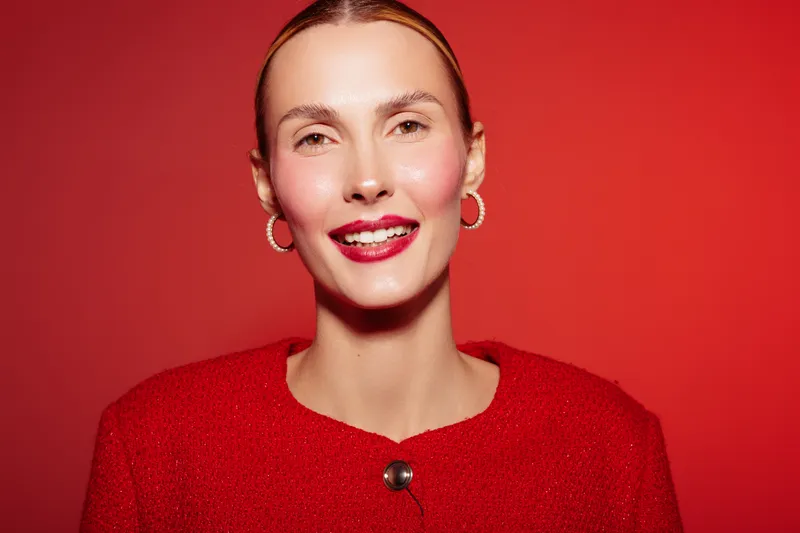 Smiling woman with red lipstick and red jacket poses on red background. Close-up portrait with hoop earrings and natural makeup. Concept of beauty, fashion, and confidence.
