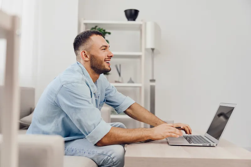 Smiling man working at home, joyful atmosphere, modern interior, casual style, laptop on table, bright colors, focused expression.