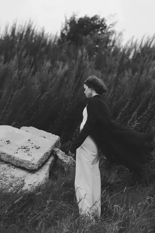 Woman in dress and flowing cape gazes toward rugged rocks in a black and white landscape, conveying mystery, elegance, and quiet solitude amid wind and grass