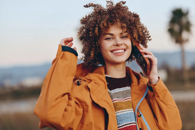 Woman with curly hair smiling outdoors lifestyle jacket travel portrait with sunny natural light She holds and adjusts a brown jacket while standing in a scenic landscape