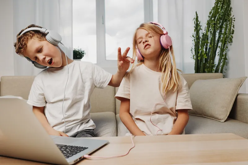 Two children wearing headphones enjoy music together at home, sitting on a sofa with a laptop nearby. Concept of childhood fun, technology, and leisure time.