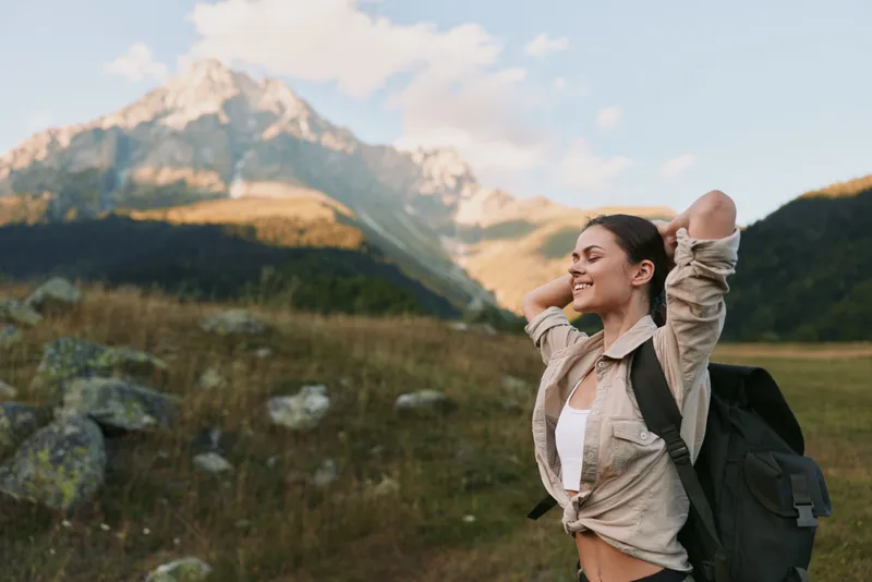 Cheerful woman outdoors, smiling with sunlit cheeks, hiking with backpack, casual jacket, amidst meadow and mountain scenery, embracing freedom and nature.