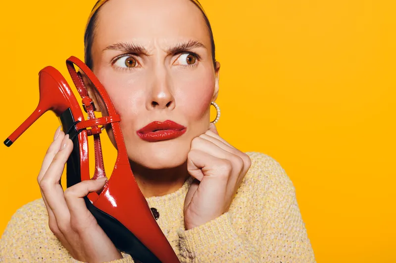 Confused woman with red lipstick holds red high heel shoe near her ear on yellow background. Concept for fashion dilemma, style choice, and unusual communication.