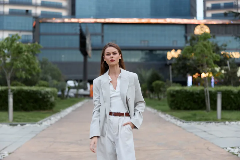 Confident woman in stylish light suit walking urban street with modern building background, casual business fashion and natural emotion