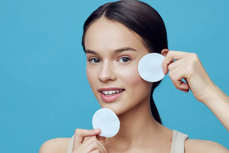 Young female model demonstrating skin care routine with cotton pads, showcasing a fresh and healthy appearance against a vibrant blue background