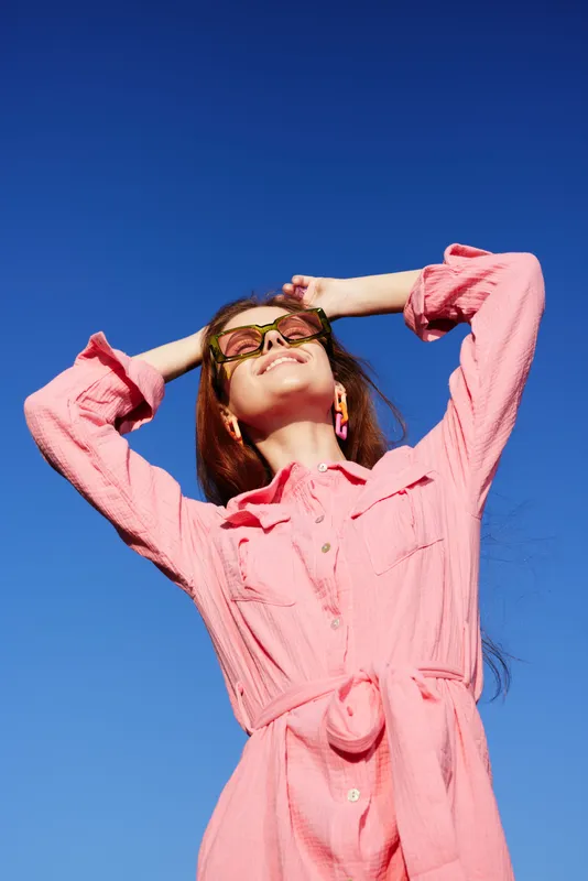 A cheerful young woman in a pink dress enjoying sunny weather against a bright blue sky, embodying summer vibes and a carefree spirit