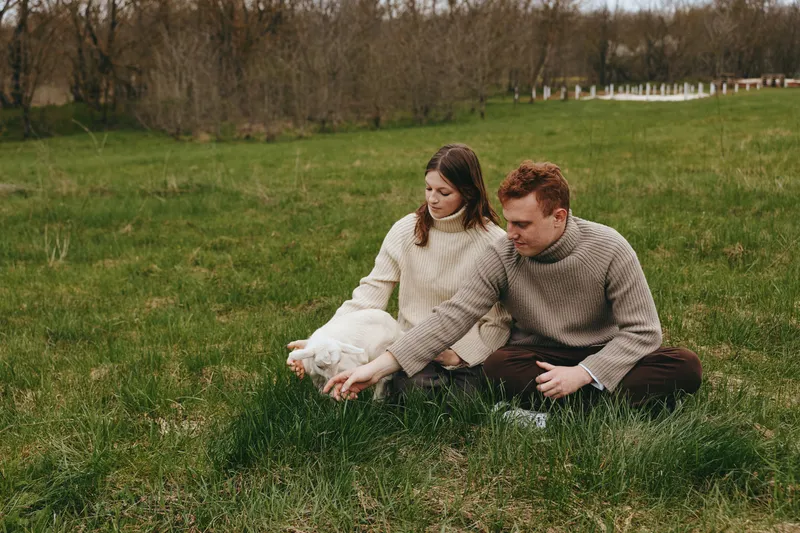 Lifestyle scene of a couple outdoors on a grassy field, smiling as they cradle a playful goat kid, cozy sweaters, natural light, gentle breeze, calm countryside atmosphere