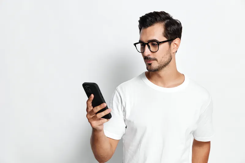 Stylish young man wearing glasses and a casual white t-shirt, gazing thoughtfully at his smartphone, showcasing a modern lifestyle against a minimalistic background.