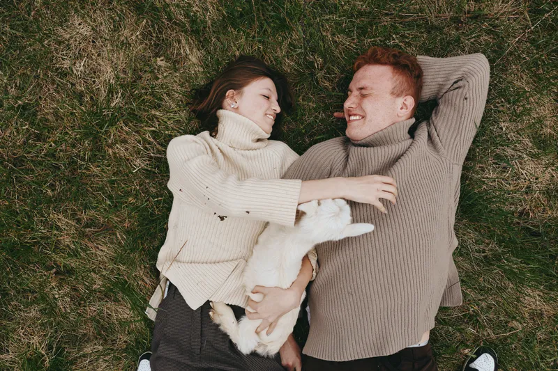 Young couple lies on the grass wearing cozy sweaters while holding a white baby goat, sharing a joyful moment in a natural lifestyle setting outdoors during autumn.