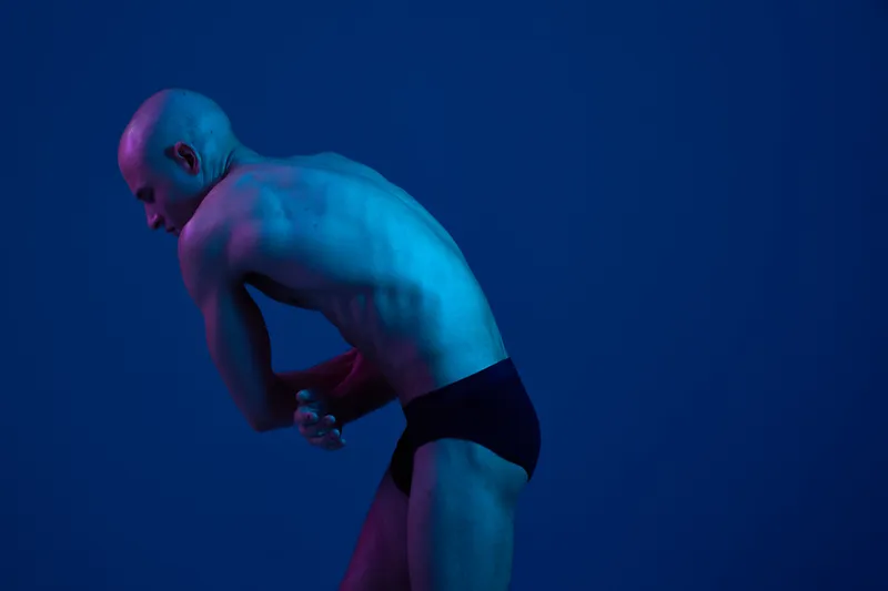 Athletic man posing in a dramatic blue and purple lighting, showcasing strong physique and artistic silhouette, emphasizing movement and form.