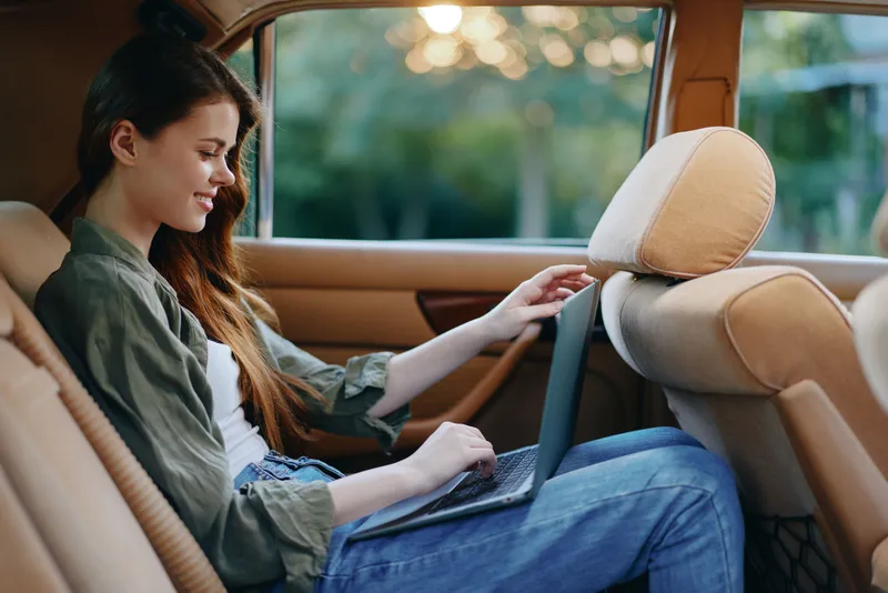 Young woman working on a laptop in a car, wearing casual attire and smiling, surrounded by a warm sunset background, emphasizing work-life balance and productivity.