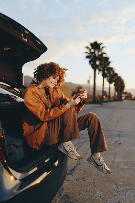 Woman lifestyle scene, woman with curly hair beside open car door on city street, holding coffee cup, wearing brown jacket and jeans, relaxed travel mood in sunset light, candid moment