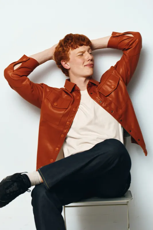 Young man with red hair sitting relaxed on stool with hands behind head wearing casual orange jacket white t shirt and dark pants against white background casual lifestyle portrait