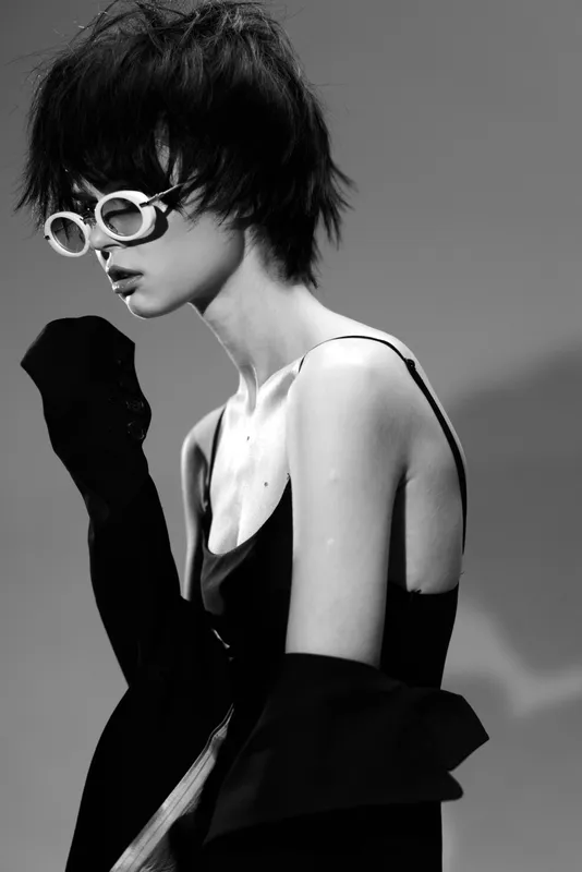 Black and white portrait of a stylish woman wearing sunglasses and a sleeveless dress, showcasing a modern and edgy fashion look.