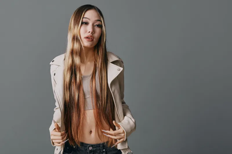 Young woman with long straight hair posing in beige jacket and cropped top, standing against grey background. Fashion portrait of confident female with casual urban style indoors.