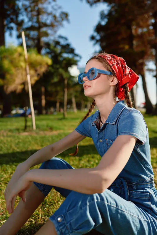 young woman wearing blue sunglasses and a red bandana sitting outdoors on green grass in warm sunlight with trees in the background in casual denim outfit