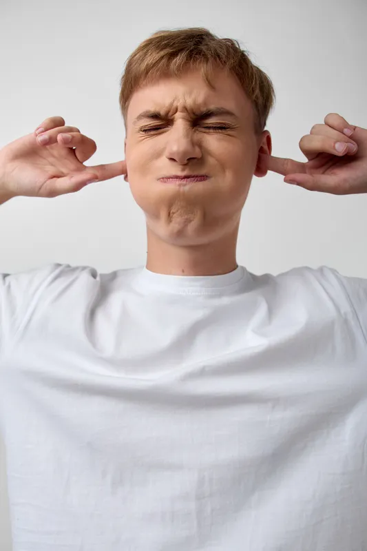 Young man in a white t shirt pinches his ears with fingers, eyes closed in discomfort, signaling loud noise, ear protection and silence in a candid moment of sensory strain