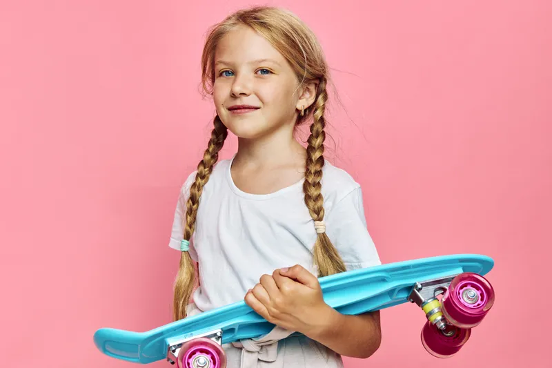 Young girl with braided hair holding a blue skateboard posing against a pink background. Casual and playful concept for youth and leisure.