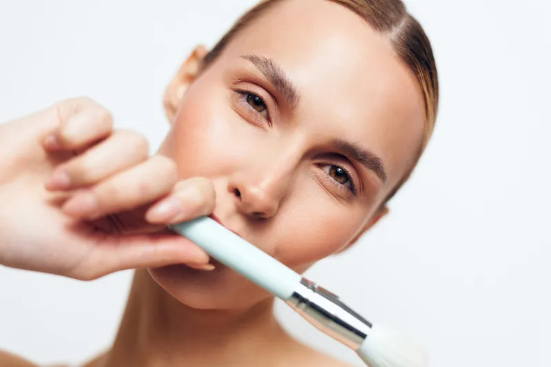 A young woman applies makeup with a large brush on her face against a white background. The close-up captures natural skin texture and focused expression. Beauty and skincare concept.
