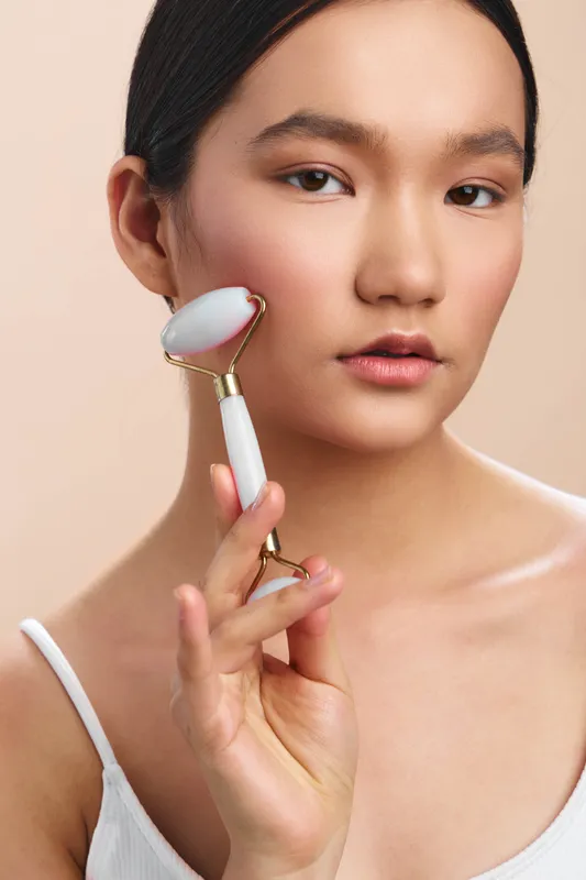 Asian woman uses a facial roller in a skincare routine, closeup portrait with serene expression, emphasizing healthy skin, glow, and selfcare in soft, focused lighting.