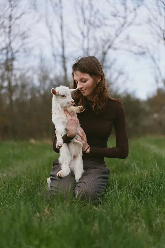 A woman in a brown top sits on green grass outdoors, cradling a small white goat kid, smiling softly as they share a tender moment. A gentle lifestyle scene with animal companionship.
