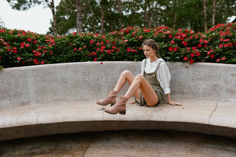 Stylish young woman in green overalls and beige boots sitting on a stone bench surrounded by vibrant flowers, showcasing a casual summer vibe