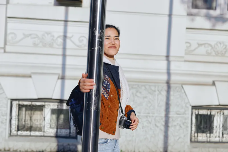 Woman in casual jacket stands beside a street pole under bright daylight, urban backdrop and confident smile, capturing a lifestyle fashion moment for street photography and everyday style