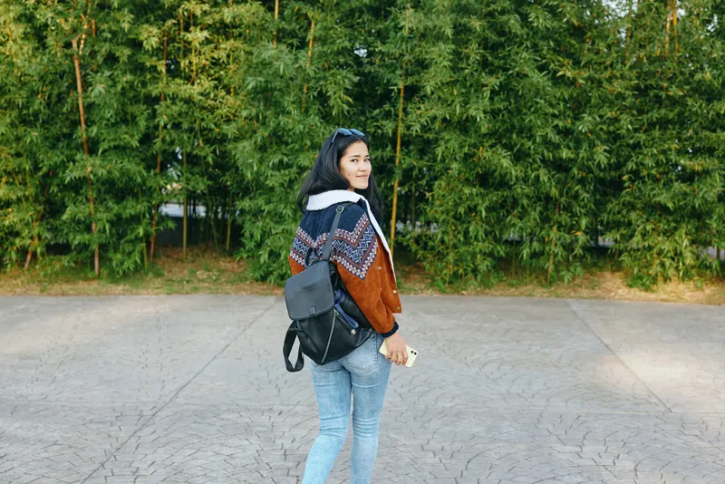 Woman with backpack walking beside a tree lined park in autumn, wearing a patterned jacket and jeans, smiling back at the camera on a paved path, outdoor scene during golden hour