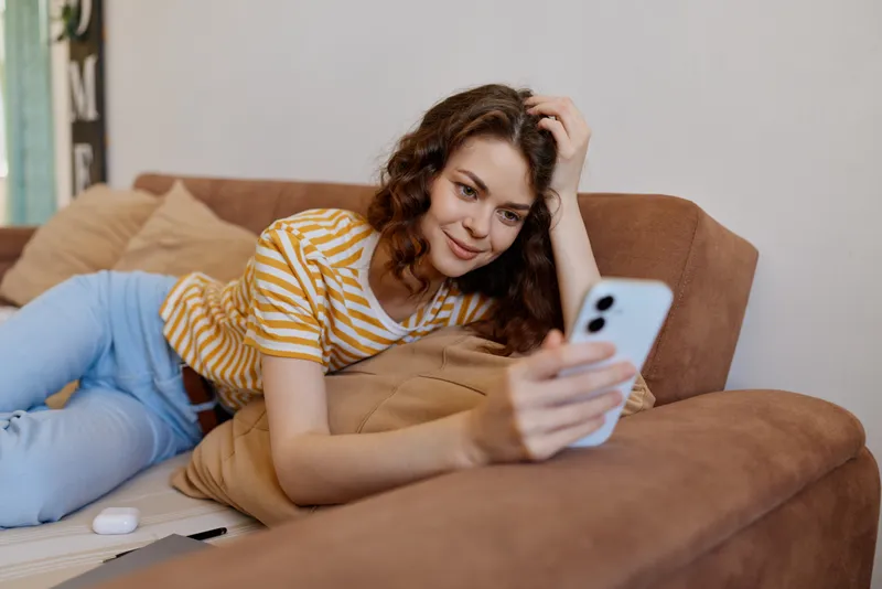 A smiling woman relaxes at home on a brown couch, browsing a smartphone. A casual lifestyle moment highlighting comfort, self expression, and everyday home life.