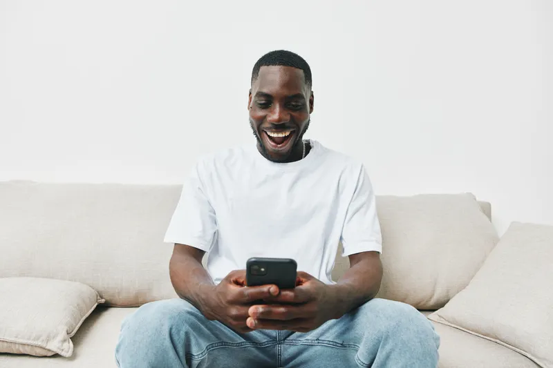 Joyful young man engaged in texting on smartphone at home, wearing casual white t shirt, expressing happiness and excitement, with a neutral background