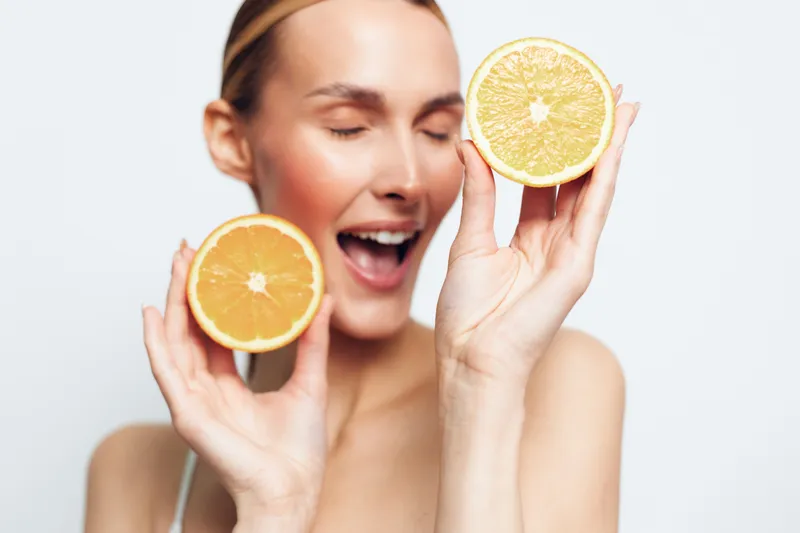 Young woman with blonde hair holds two halves of a grapefruit near her face while smiling with closed eyes against a white background.