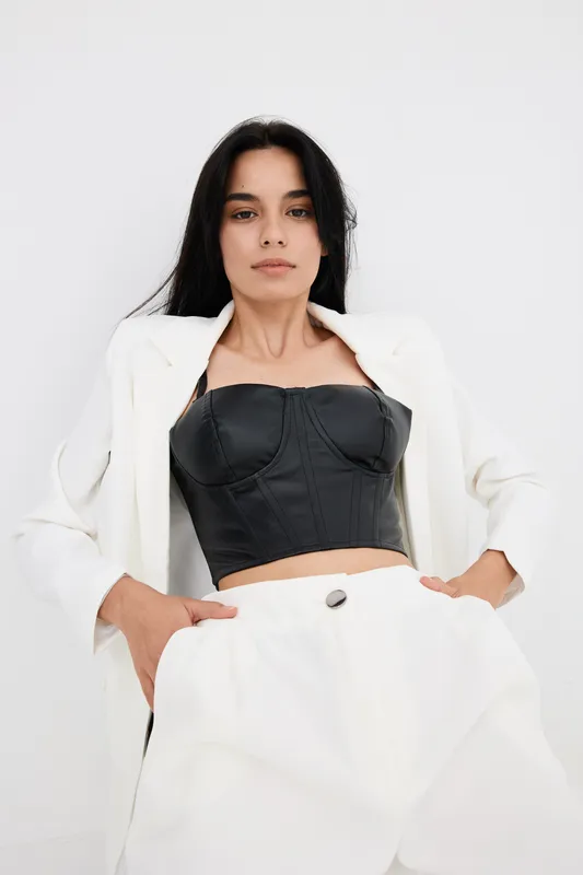 Woman with long black hair wearing black leather corset and white oversized suit posing confidently against plain white background, modern fashion style.