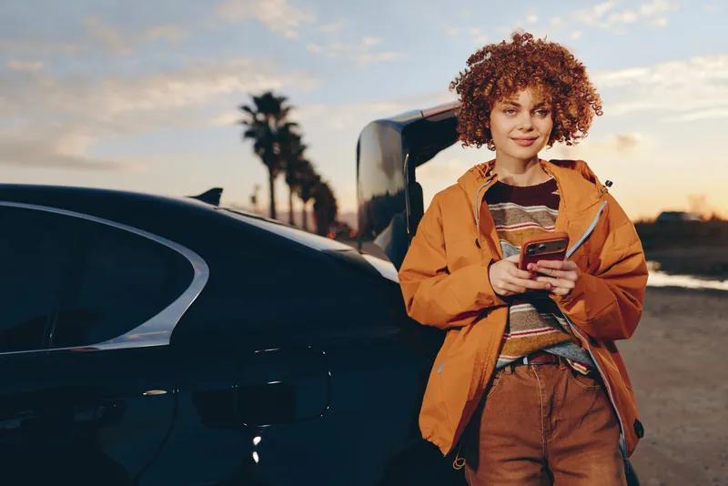 Cheerful woman lifestyle outdoors near car, holding smartphone, wearing orange jacket, curly hair, walking along road at sunset, relaxed posture, palm trees in background, warm soft light